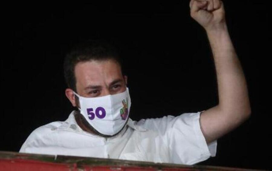 Guilherme Boulos na sacada de sua casa, de máscara, ergue o braço com o punho cerrado
