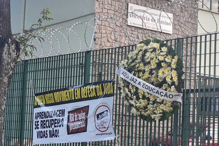 Foto de uma faixa da Apeoesp pedindo respeito à vida e, ao seu lado, uma coroa de flores dizendo "Aqui jaz a Educação", nas grades dos portões de entrada da Diretoria de Ensino Centro, na Casa Verde.