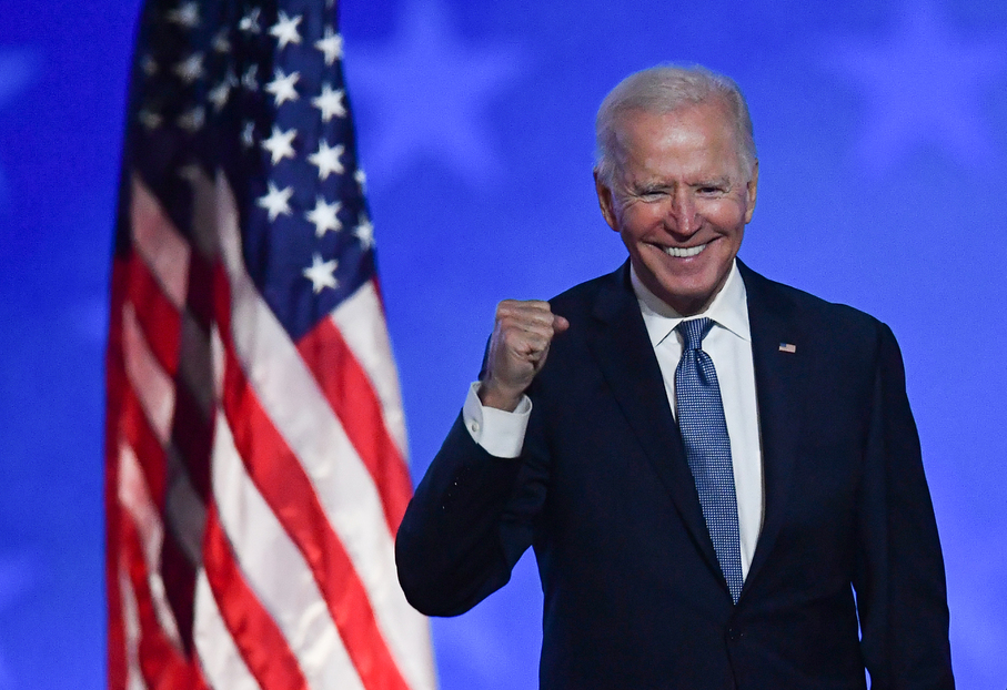 Joe Biden comemora sua vitória nas eleições. Sorrindo e com o braça meio levantado e punhos cerrados com a bandeira dos EUA atrás