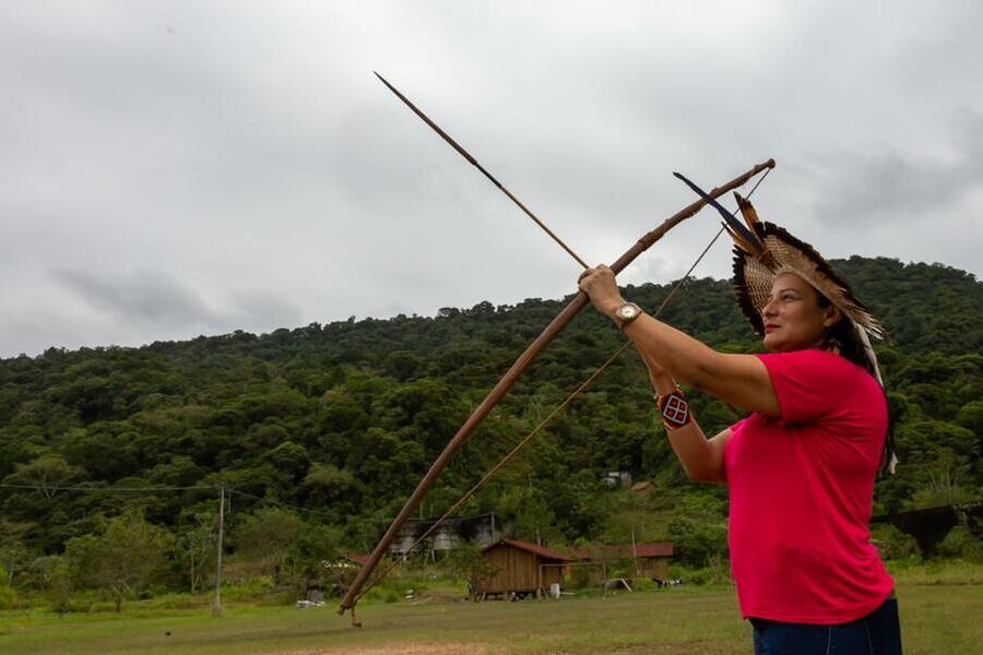 Vereadora Juliana Cardoso usando um cocar, prepara o arco para atirar uma flecha