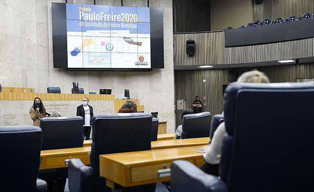 Plano parcial da mesa de trabalhos da Câmara, destacando painel com a inscrição Prêmio Paulo Freire, 2020