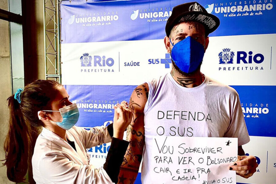 Tico Santa Cruz porta um cartaz enquanto a atendente do SUS aplica a vacina contra a Covid-19