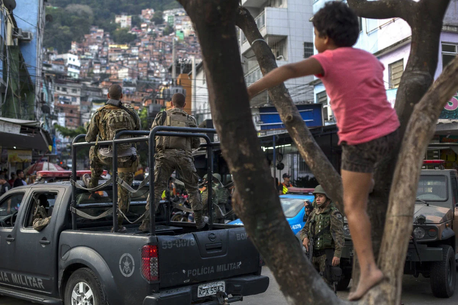 Uma criança, em cima da árvore, observa a tropa da PM entrando num carro blindado na favela