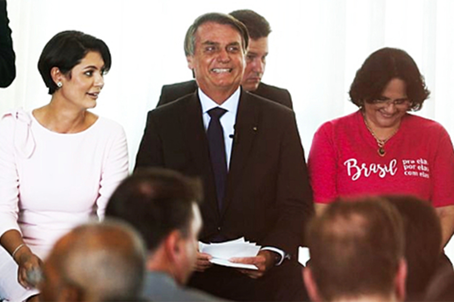 Bolsonaro ri, tendo sua mulher, Michele Bolsonaro do seu lado direito, e a ex-ministra Damares Alves do seu lado esquerido
