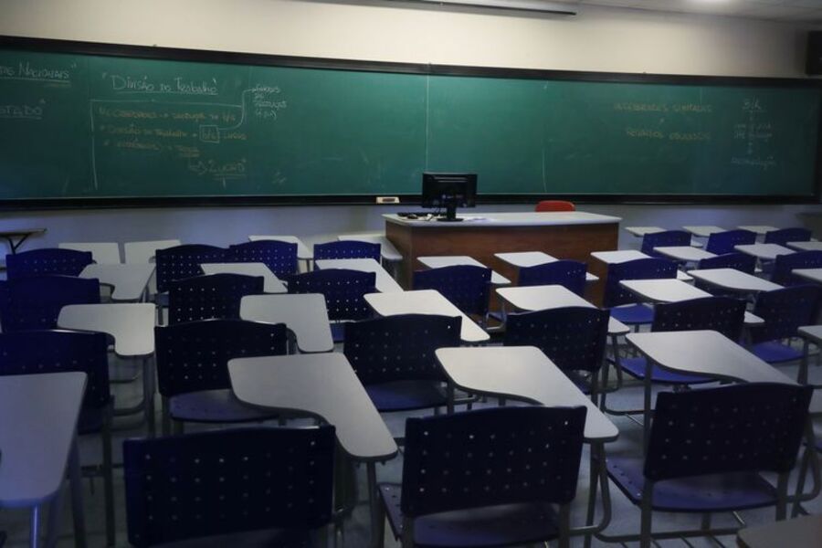 Foto do interior de uma sala de aula vazia. Observa-se a quantidade enorme de carteiras na sala