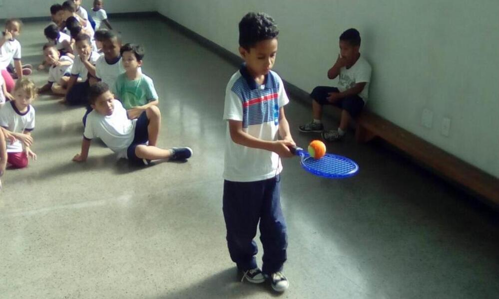 Criança, em atividade física na escola, brinca de equilibrar uma bola na raquete de tênis