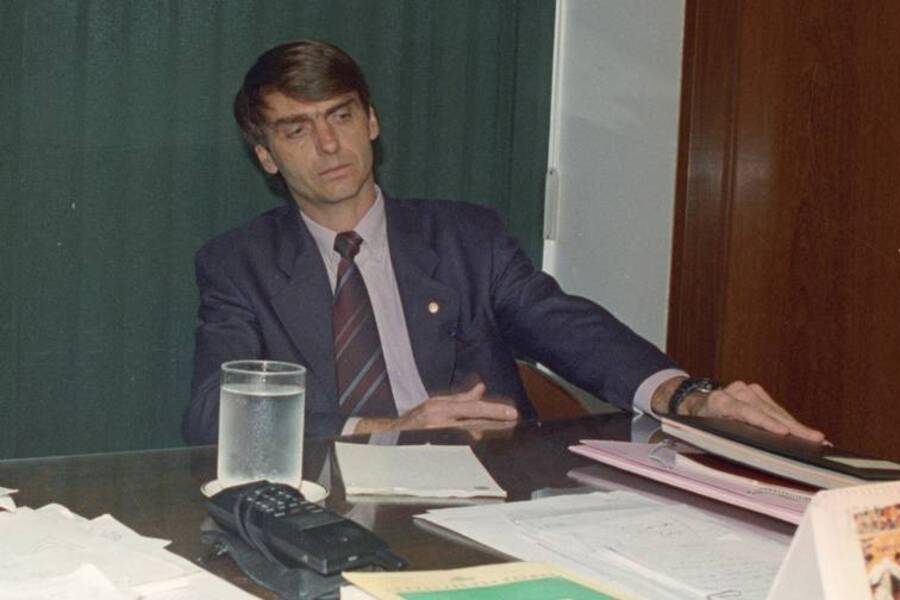 Foto antiga do então deputado federal Jair Bolsonaro, que está sentado à mesa mexendo em alguns papéis.