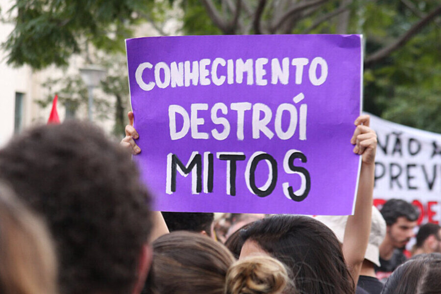 Foto de uma professora que ergue um cartaz com a frase "Conhecimento destrói Mitos"
