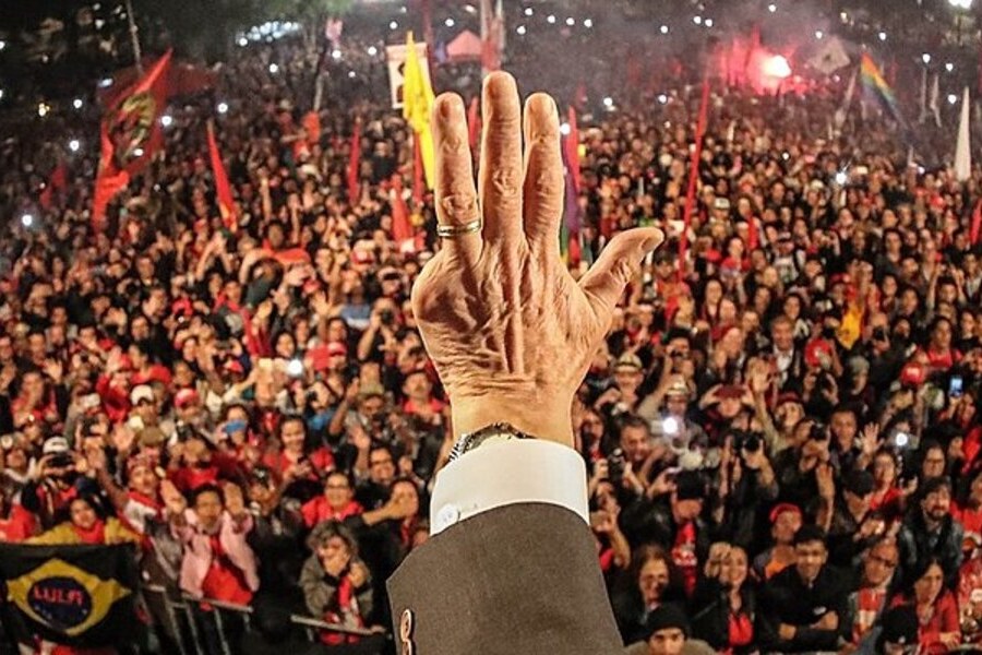 Multidão, de frente para a câmara, observa mão estendida, em primeiro plano, de Lula.