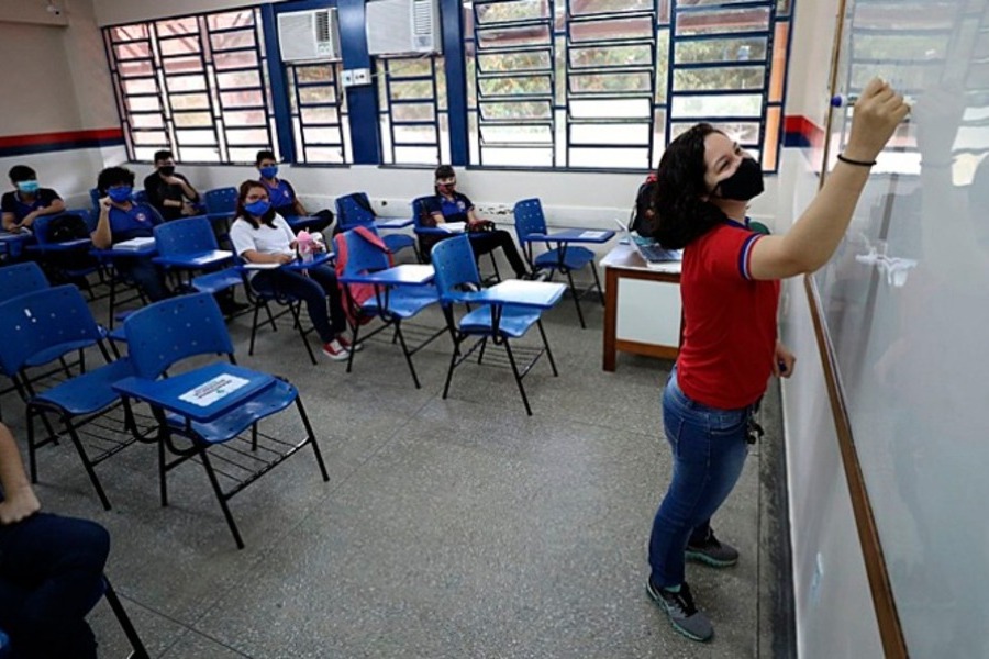 Foto ilustrativa de professora, em aula, escrevendo na lousa, enquanto poucos alunos assistem a aula.