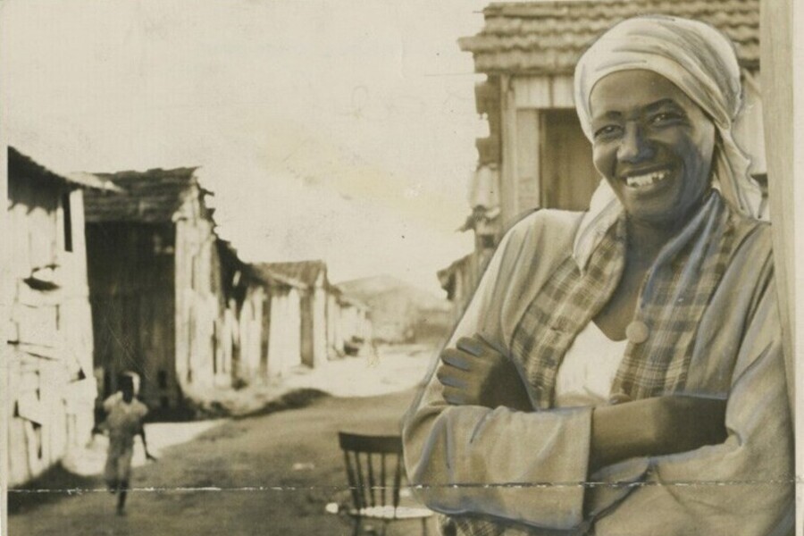 Foto de Carolina Maria de Jesus sorrindo, de braços cruzados e encostada na parede de sua casa, na ex-favela do Canindé