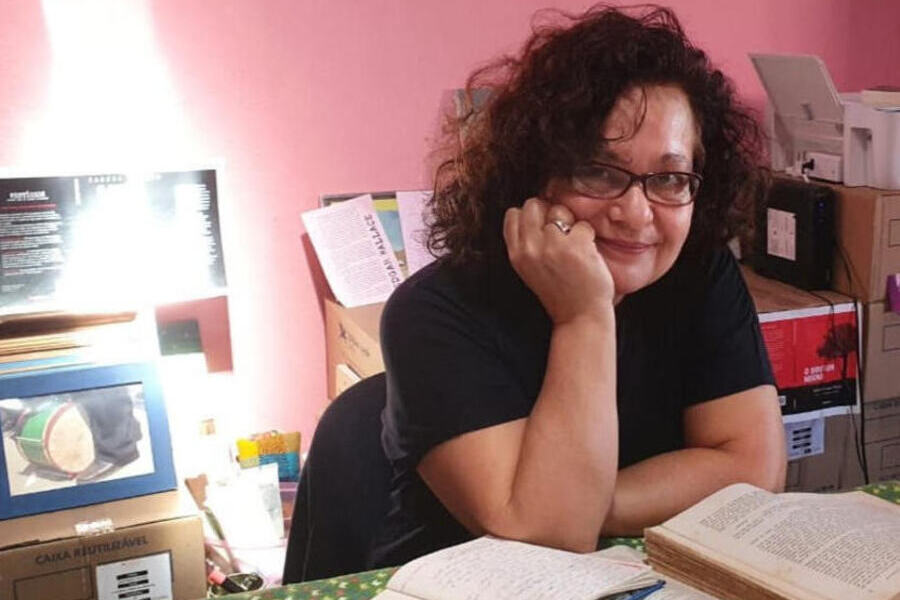 Foto da escritora Sandra Abrano, que está sentada diante de uma mesa cheia de livros e anotações. Está com uma das mãos no queixo, segurando a cabeça
