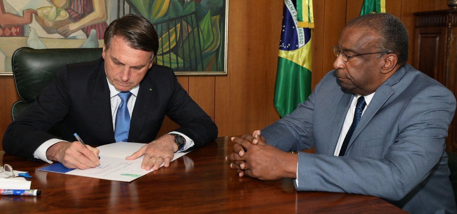 Foto do presidente Bolsonaro e do ex-ministro Decotelli sentados à mesa. Bolsonaro está à esquerda e Decotelli à direita. Bolsonaro está assinando a nomeação do ex-ministro na sala presidencial