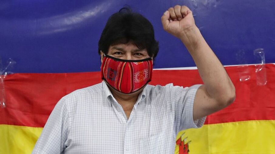 Evo Morales, de máscara, ergue o braço com o punho fechado. Atrás dele, a bandeira da Bolívia