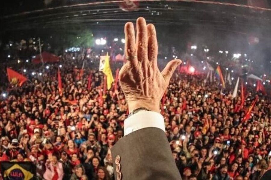Foto da mão esquerda de Lula estendida. Ao fundo, uma multidão o escuta