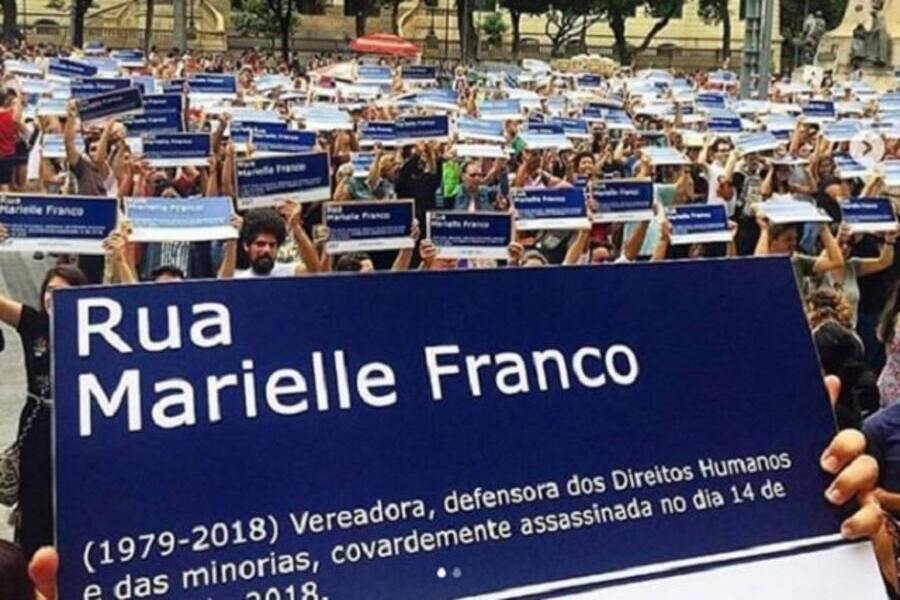 Foto de manifestação em que centenas de pessoas erguem uma placa de rua com o nome de Marielle Franco