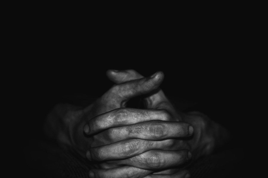 Mãos com os dedos entrecruzados de homem destaca-se num cenário absolutamente escuro.