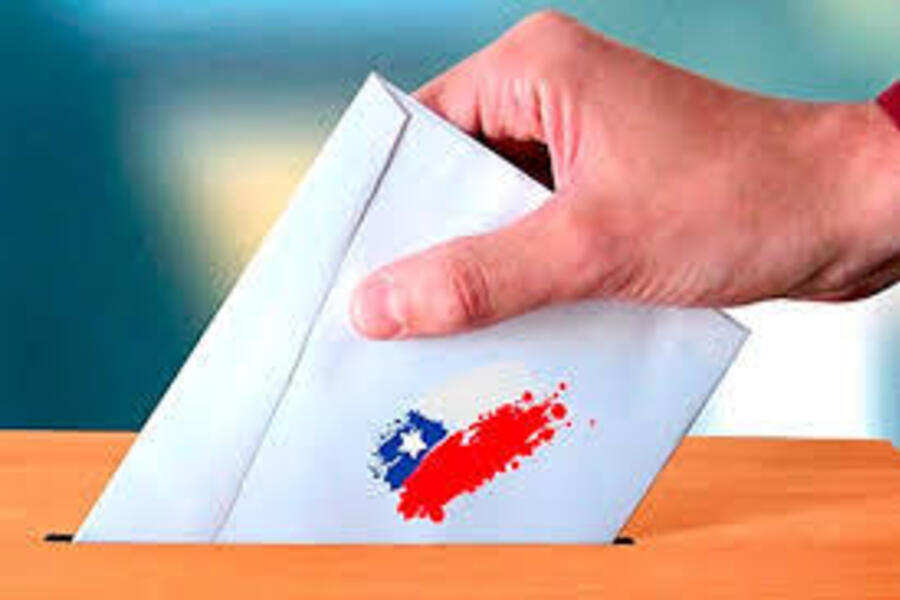 Mão masculina introduz envelope de votação em urna. Nesse envelope está estampado a bandeira estilizada do Chile