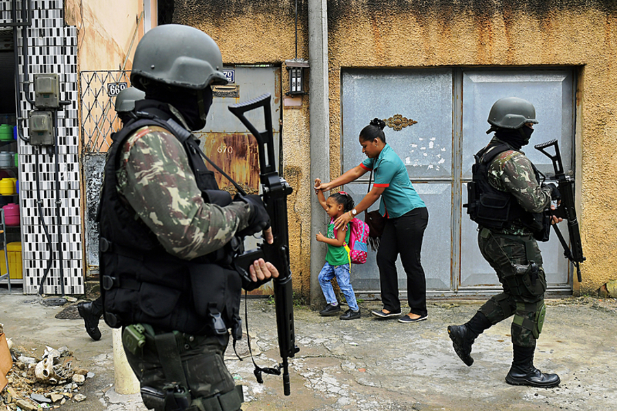 Soldados da PM carioca entram em comunidade. No destaque dois policiais fortemente armados. Ao fundo, mãe protege sua filha 