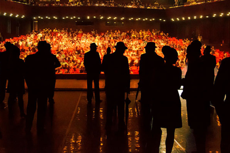 Foto tirada dos bastidores, mostra atores em palco e, ao fundo, a plateia