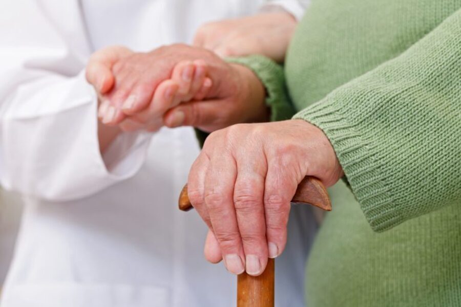 Foto de close de um cuidador segurando a mão de uma pessoa idosa que ampare-se numa bengala