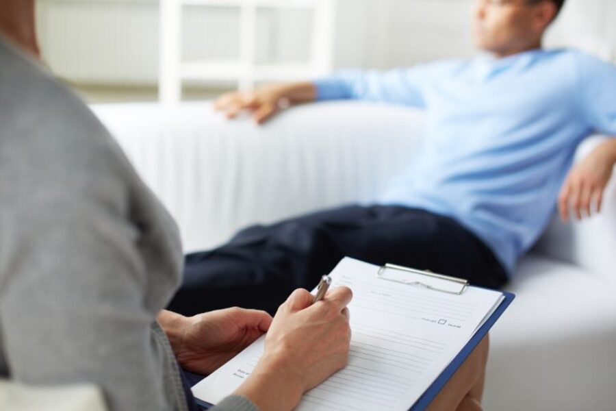 Paciente sentado a um sofá em sessão com seu psicólogo que anota o que diz em um caderno. 