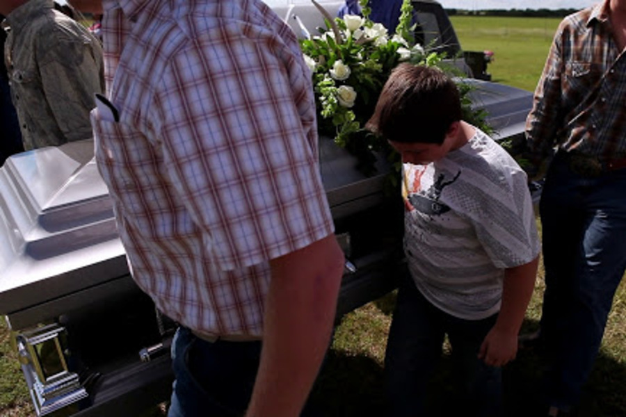 Três pessoas carregam um caixão para a sepultura. Uma dessas pessoas é uma criança.