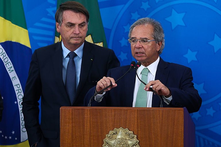 Ministro Paulo Guedes fala ao microfone com Bolsonaro bem atrás dele