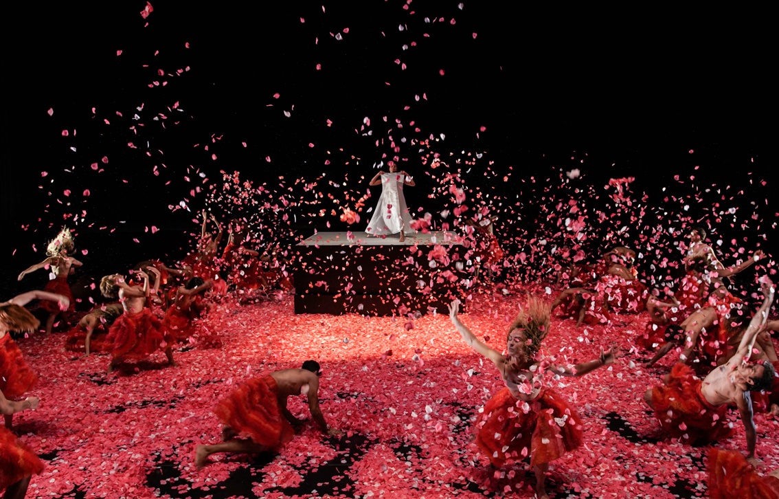Foto do espetáculo visto da plateia. Dançarinos em ação, luz vermelha sobre cenário também em vermelho. Papéis picados caem como se chovesse no palco