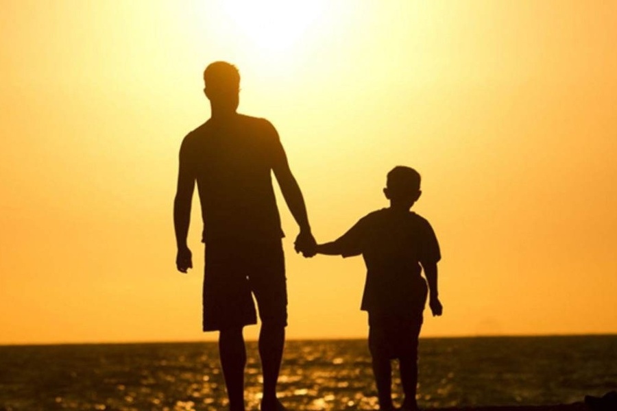 Foto de pai de mãos dadas com o filho contemplando à beira-mar, o pôr do Sol