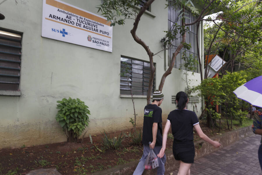 Foto parcial do prédio do AE do Tucuruvi, onde se vê a placa de identificação do Ambulatório preso à parede. Duas pessoas passam em frente.