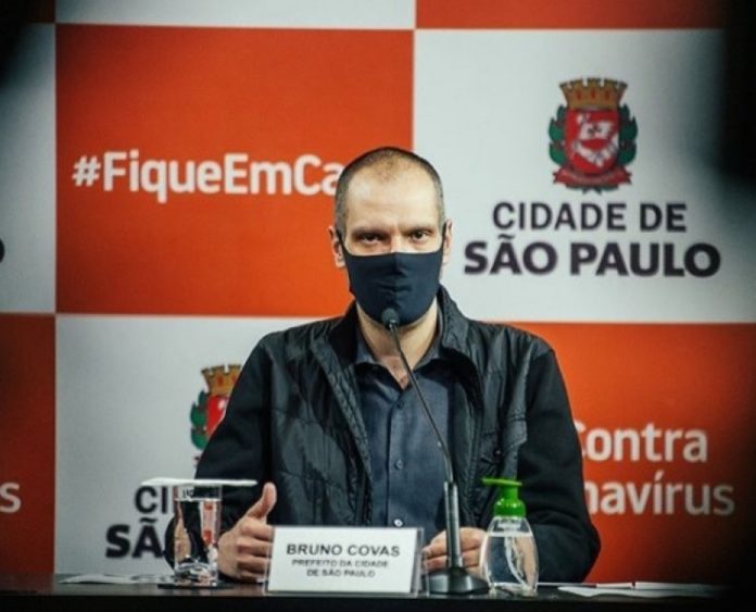 Foto do Prefeito sentado de máscara. Ao fundo logo da Prefeitura de São Paulo