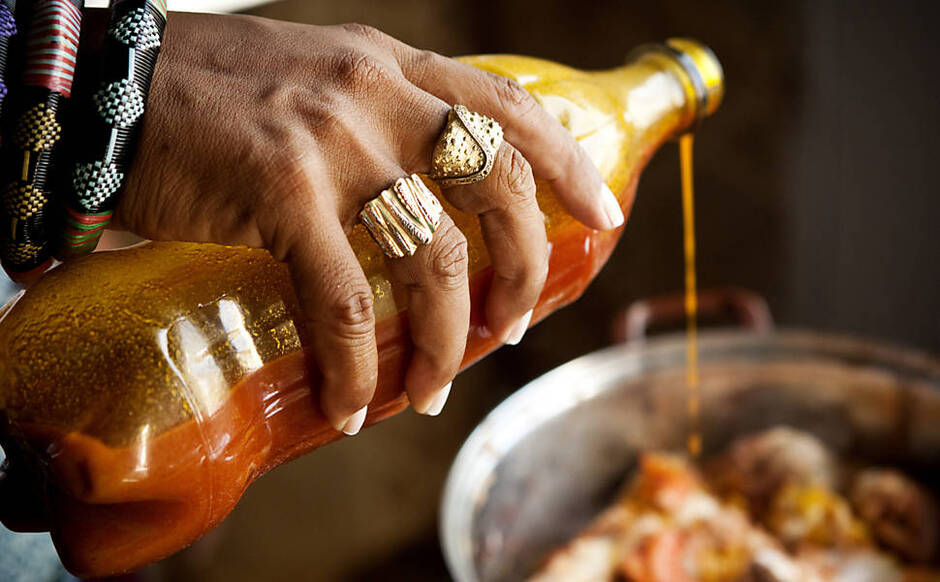 Foto mostra a mão de uma cozinheira negra cheia de anéis e pulseiras africanas segurando e despejando uma garrafa de azeite-de-dendê em uma panela